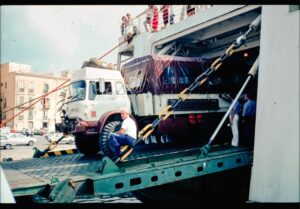A car being loaded onto a multi-vehicle open carrier truck on a suburban street with trees and driveways nearby.