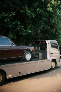 A large car carrier truck parked on a quiet suburban street, with a sedan being loaded by workers nearby.